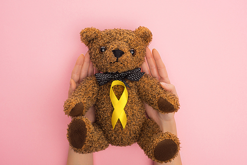 Cropped view of woman holding teddy bear with yellow ribbon on pink background, international childhood cancer day concept
