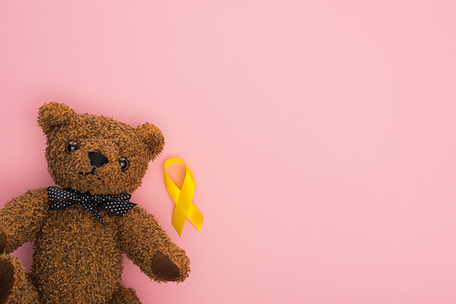 Top view of yellow ribbon near teddy bear on pink background, international childhood cancer day concept