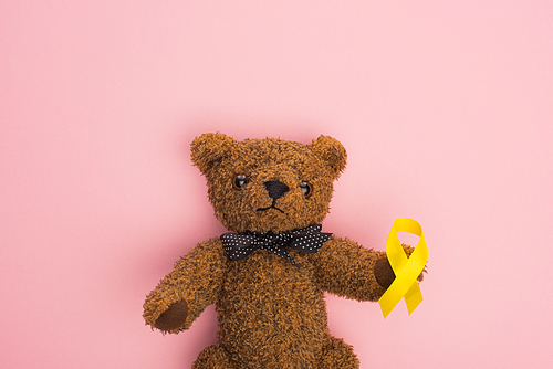 Top view of yellow ribbon on brown teddy bear with bow on pink, international childhood cancer day concept