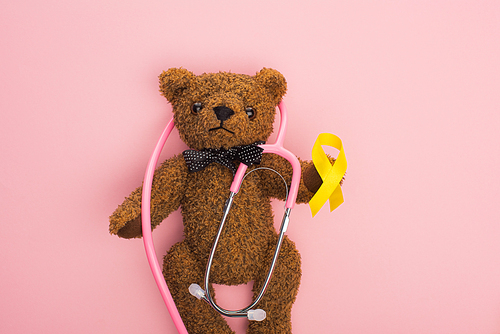 Top view of stethoscope with yellow ribbon on brown teddy bear on pink background, international childhood cancer day concept
