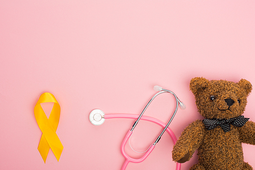 Top view of yellow ribbon near stethoscope and teddy bear on pink background, international childhood cancer day concept