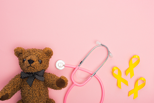Top view of yellow ribbons next to stethoscope and teddy bear on pink background, international childhood cancer day concept