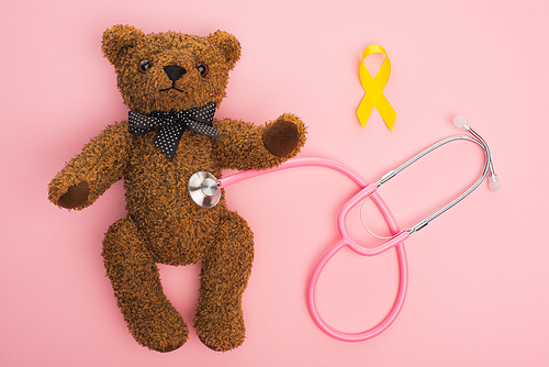 Top view of yellow ribbon and stethoscope connected with teddy bear on pink background, international childhood cancer day concept