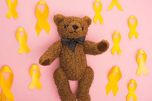 Top view of yellow ribbons around teddy bear on pink background, international childhood cancer day concept
