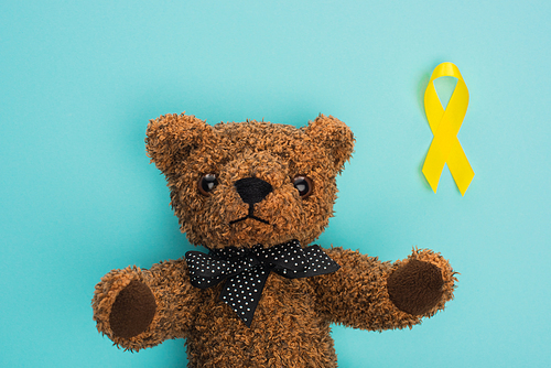 Top view of yellow ribbon next to teddy bear on blue background, international childhood cancer day concept