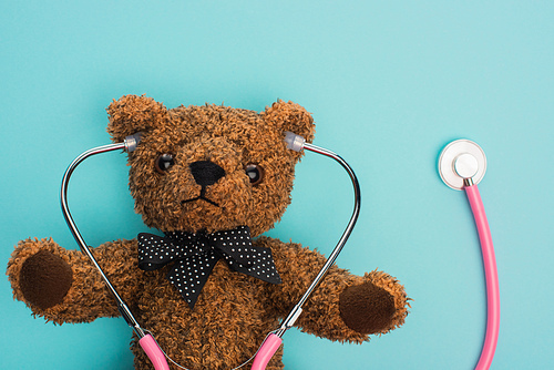 Top view of brown teddy bear with pink stethoscope on blue background, international childhood cancer day concept