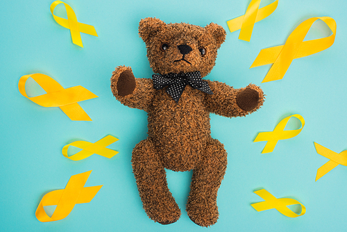Top view of yellow ribbons and brown teddy bear with bow on blue background, international childhood cancer day concept
