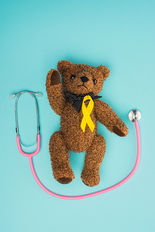 Top view of yellow ribbon on brown teddy bear near stethoscope on blue background, international childhood cancer day concept