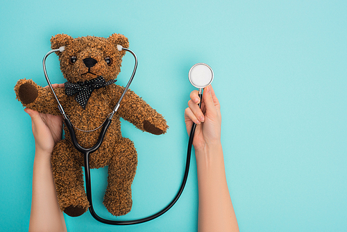 Cropped view of woman holding teddy bear with stethoscope on blue background, international childhood cancer day concept