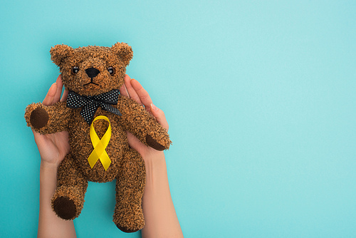 Cropped view of woman holding teddy bear with yellow ribbon on blue background, international childhood cancer day concept