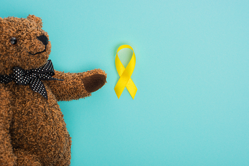 Top view of teddy bear with bow and yellow ribbon on blue background, international childhood cancer day concept