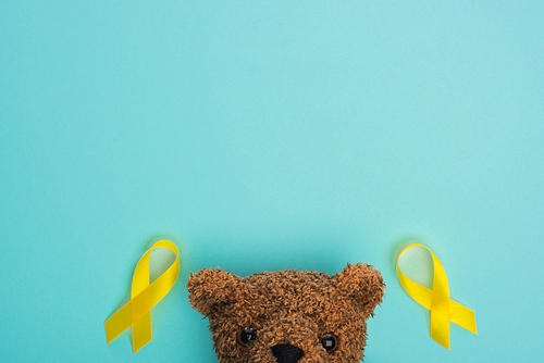 Top view of yellow awareness ribbons and brown teddy bear on blue background, international childhood cancer day concept