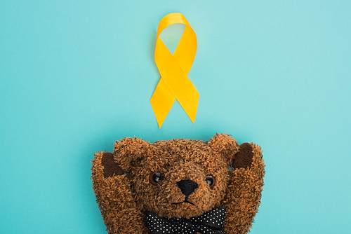 Top view of yellow ribbon with brown teddy bear on blue background, international childhood cancer day concept