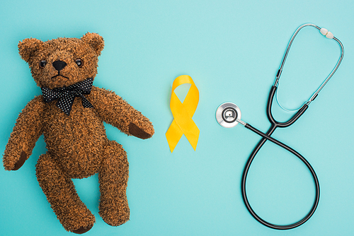 Top view of yellow awareness ribbon, teddy bear near stethoscope on blue background, international childhood cancer day concept