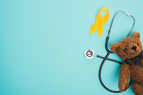 Top view of yellow ribbon, toy and stethoscope on blue background, international childhood cancer day concept