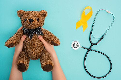 Cropped view of woman holding teddy bear near yellow awareness ribbon and stethoscope on blue background, international childhood cancer day concept