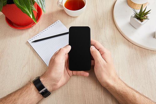 cropped view of man holding smartphone near green plants, cup of tea and blank notebook with pencil on wooden surface