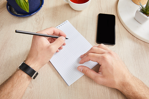 cropped view of man writing in notebook near smartphone on wooden surface