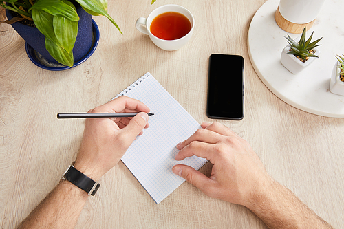 cropped view of man writing in notebook near green plants, cup of tea and smartphone on wooden surface
