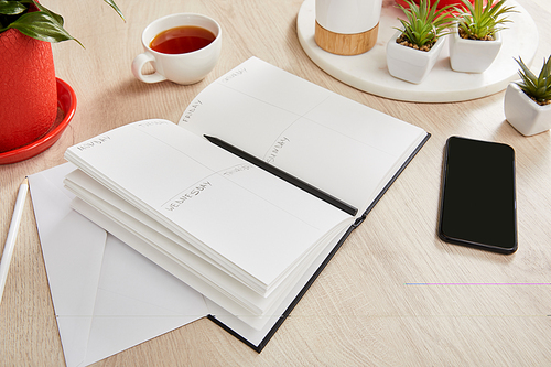 green plants, cup of tea, envelope, notebook with pencils near smartphone on wooden surface
