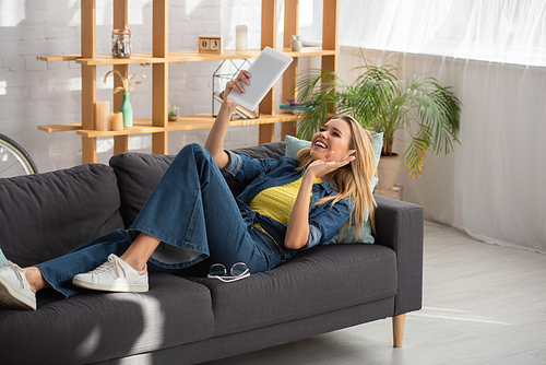 Smiling blonde woman with waving hand looking at digital tablet while lying on couch at home