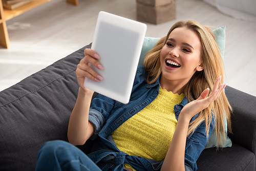 Cheerful blonde woman with waving hand looking at digital tablet while lying on couch on blurred background