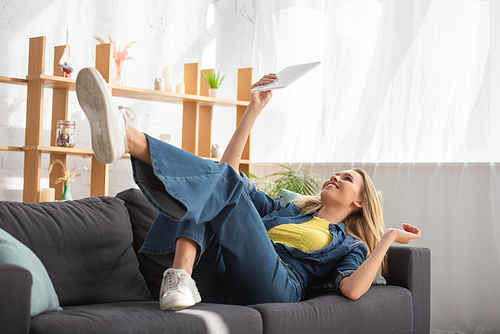 Young blonde woman looking at digital tablet while lying on couch at home on blurred background