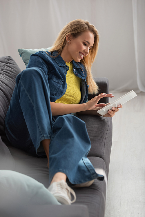 Smiling blonde woman using digital tablet while lying on couch at home on blurred foreground
