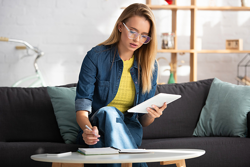 Young blonde woman with digital tablet writing in notebook while sitting on couch on blurred background