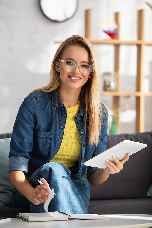 Cheerful young woman in eyeglasses  while flipping through notebook at home on blurred background