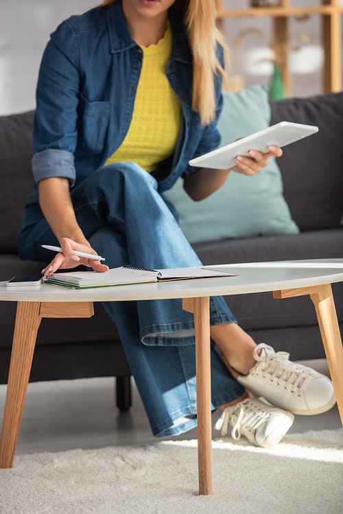 Cropped view of young woman with pen and digital tablet flipping through notebook at home on blurred background