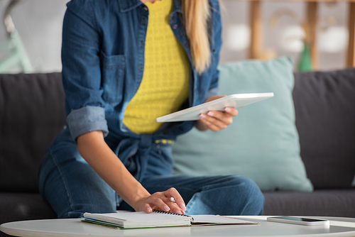 Cropped view of young woman with digital tablet sitting near notebook at home on blurred background
