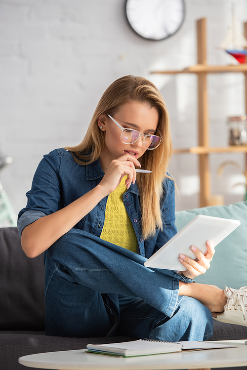 Thoughtful blonde woman with pen looking at digital tablet while sitting on couch on blurred background