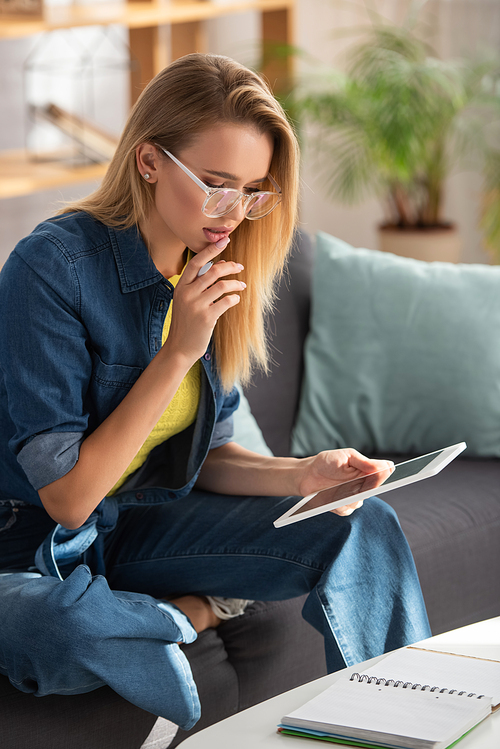 Thoughtful young blonde woman looking at digital tablet while sitting on couch at home on blurred background