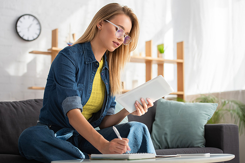 Young blonde woman with digital tablet writing in notebook while sitting on couch at home on blurred background