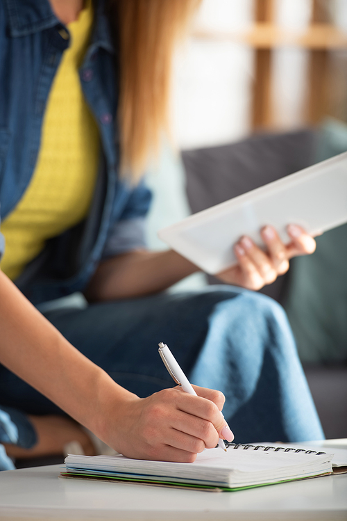 Cropped view of young woman with digital tablet writing in notebook at home on blurred background