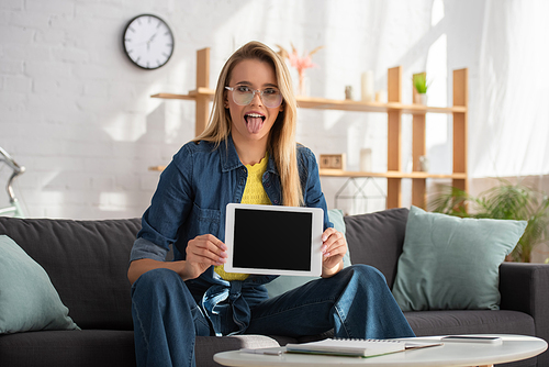 Young blonde woman with sticking out tongue  while showing digital tablet at home on blurred background