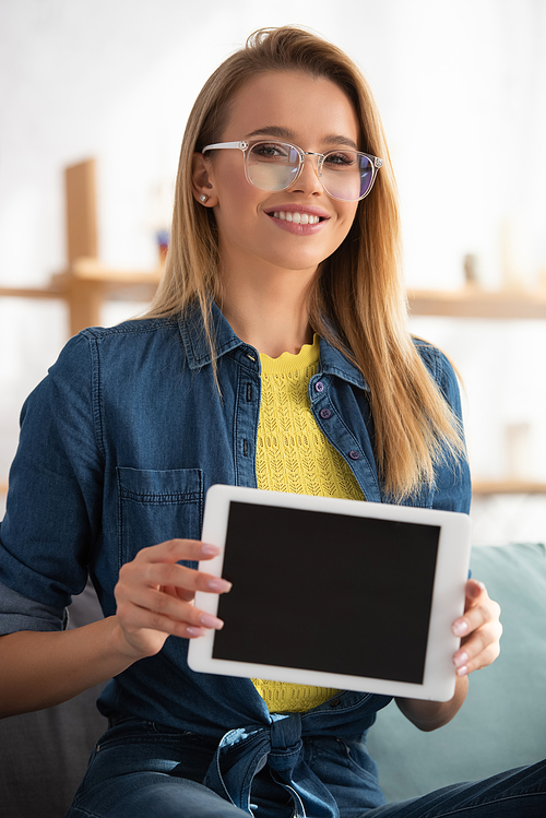 Smiling blonde woman in eyeglasses  while showing digital tablet on blurred background