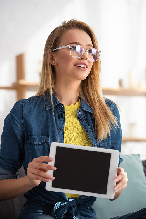 Positive blonde woman in eyeglasses looking away while showing digital tablet on blurred background