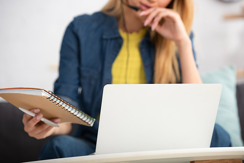 Cropped view of young woman holding notebook and pen near laptop at home on blurred background