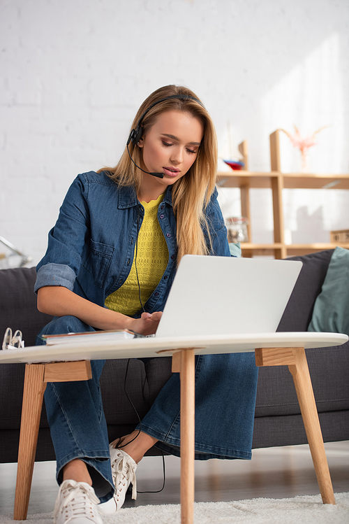 Young blonde woman in headset typing on laptop while sitting on couch near coffee table on blurred background