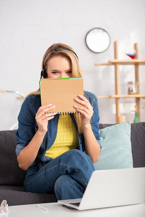 Young woman in headset  and covering face with notebook while sitting on couch on blurred background