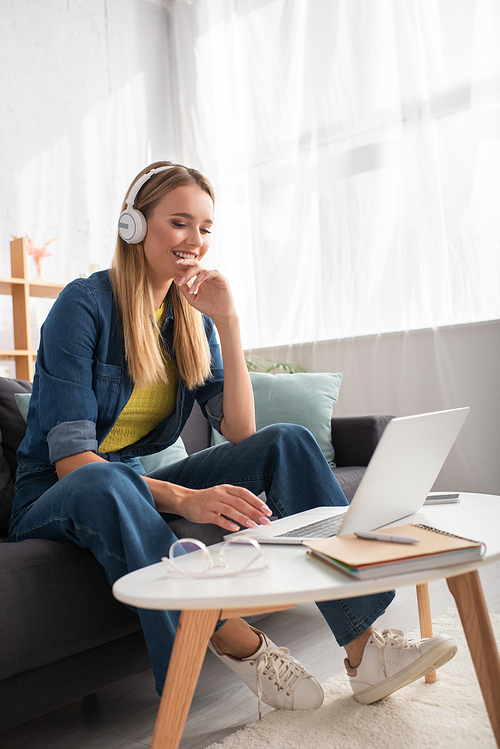 Happy young woman in headphones looking at laptop while sitting on couch near coffee table on blurred background