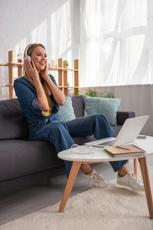 Full length of cheerful blonde woman in headphones laughing while sitting on couch near devices on coffee table