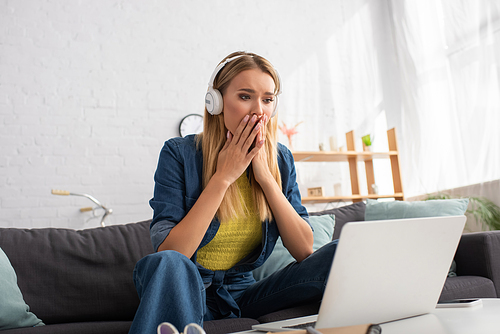 Frightened blonde woman in headphones looking at computer while sitting on couch at home on blurred background