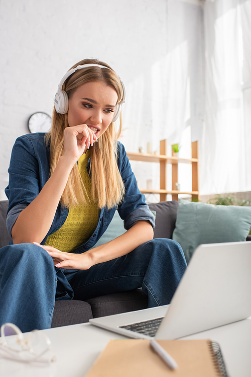 Worried young woman in headphones looking at laptop while sitting on couch on blurred foreground
