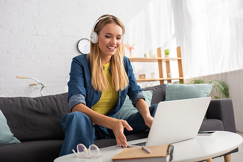 Cheerful blonde woman in headphones looking at laptop while sitting on couch near coffee table on blurred background