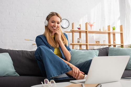 Happy blonde woman in headphones looking at laptop while sitting on couch at home