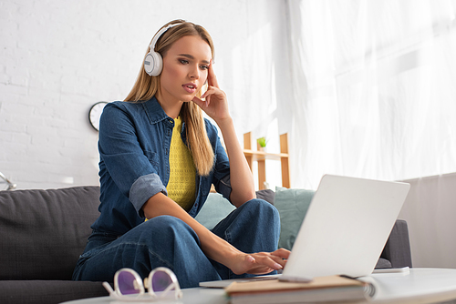 Thoughtful young woman in headphones using laptop while sitting on couch at home on blurred foreground