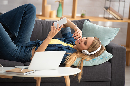 Smiling blonde woman in headphones looking at smartphone while lying on couch near coffee table on blurred background